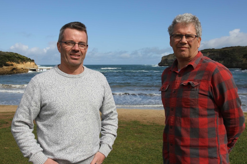 Two men stand in front of a beach.