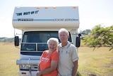 An older couple stand in front of their motor home with arms around each other smiling.