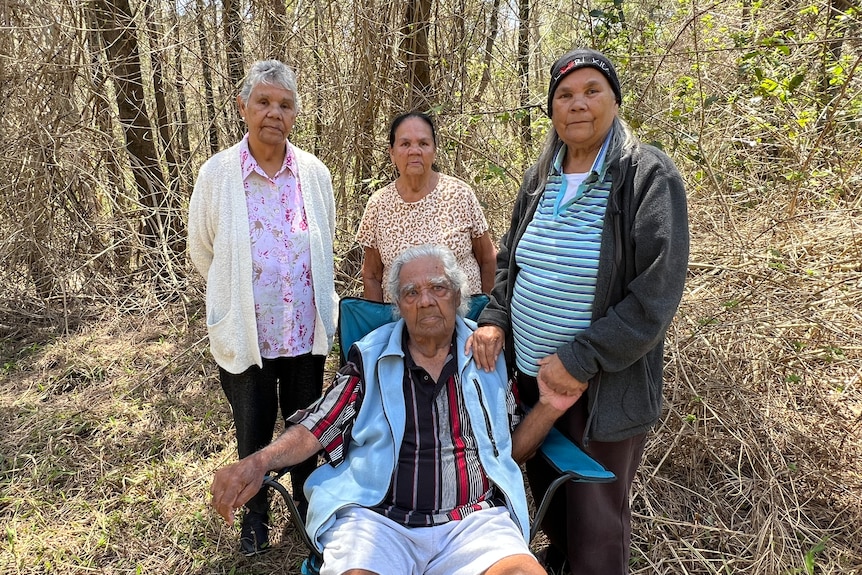 Elderly Aboriginal man sitting on a chair in an overgrown bush area with three elderly women standing behind him
