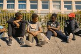Five young black boys sit in a row on pavement, against a metal fence, talking.