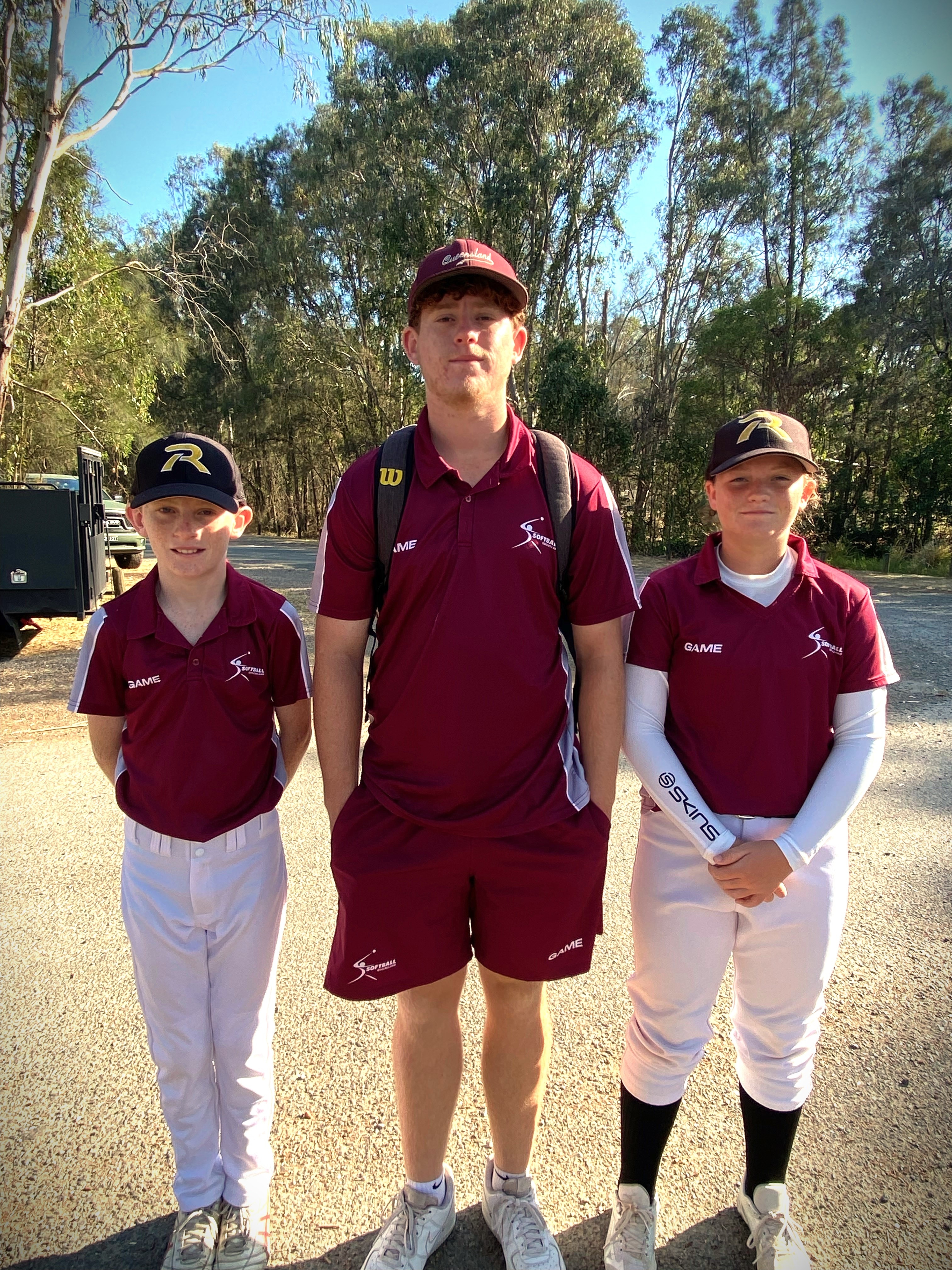 Three children standing together in maroon shirts and wearing softball caps with trees in the background.