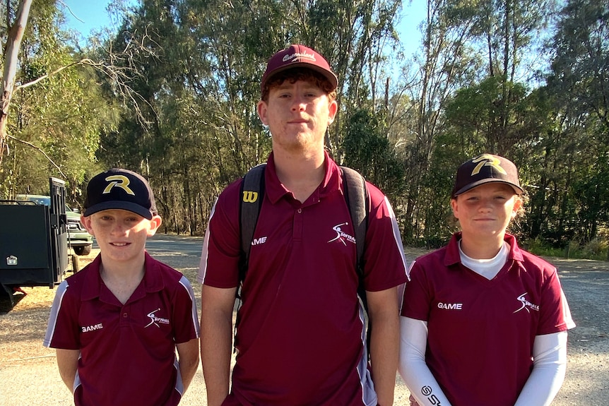 Three children standing together in maroon shirts and wearing softball caps with trees in the background.