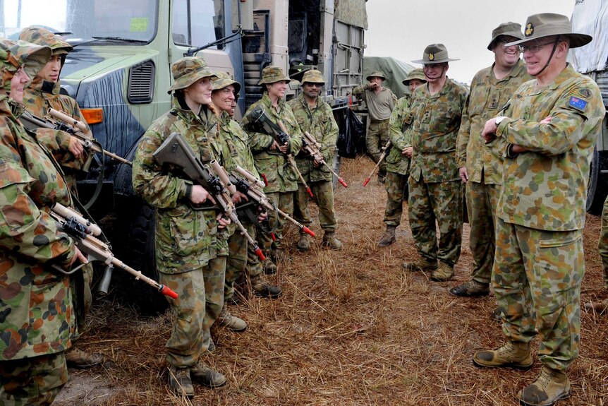 A group of adults wearing camouflage and holding firearms with orange tips stand around casually on hay smiling