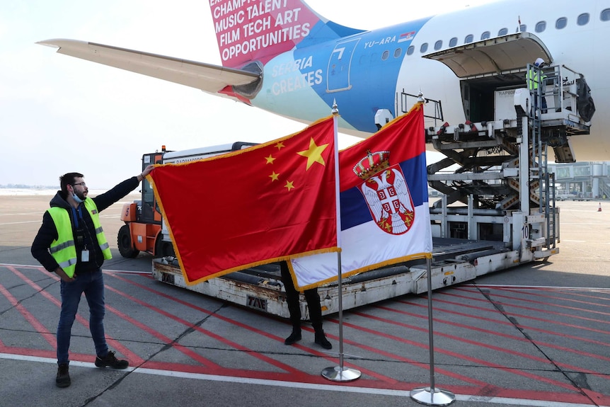 A man holds China's flag next to Serbia's flag as Sinopharm COVID-19 vaccines are offloaded from the cargo door of a jet.