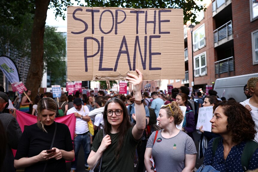 A woman in a crowd holding a sign reading "Stop the Plane" 