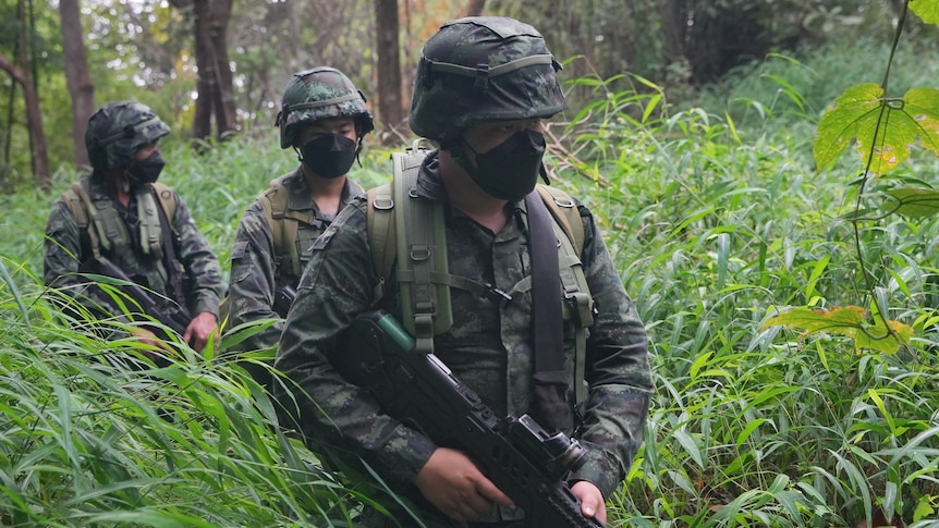Three soldiers in camouflage gear make their way through dense, tall grass in a jungle clearing