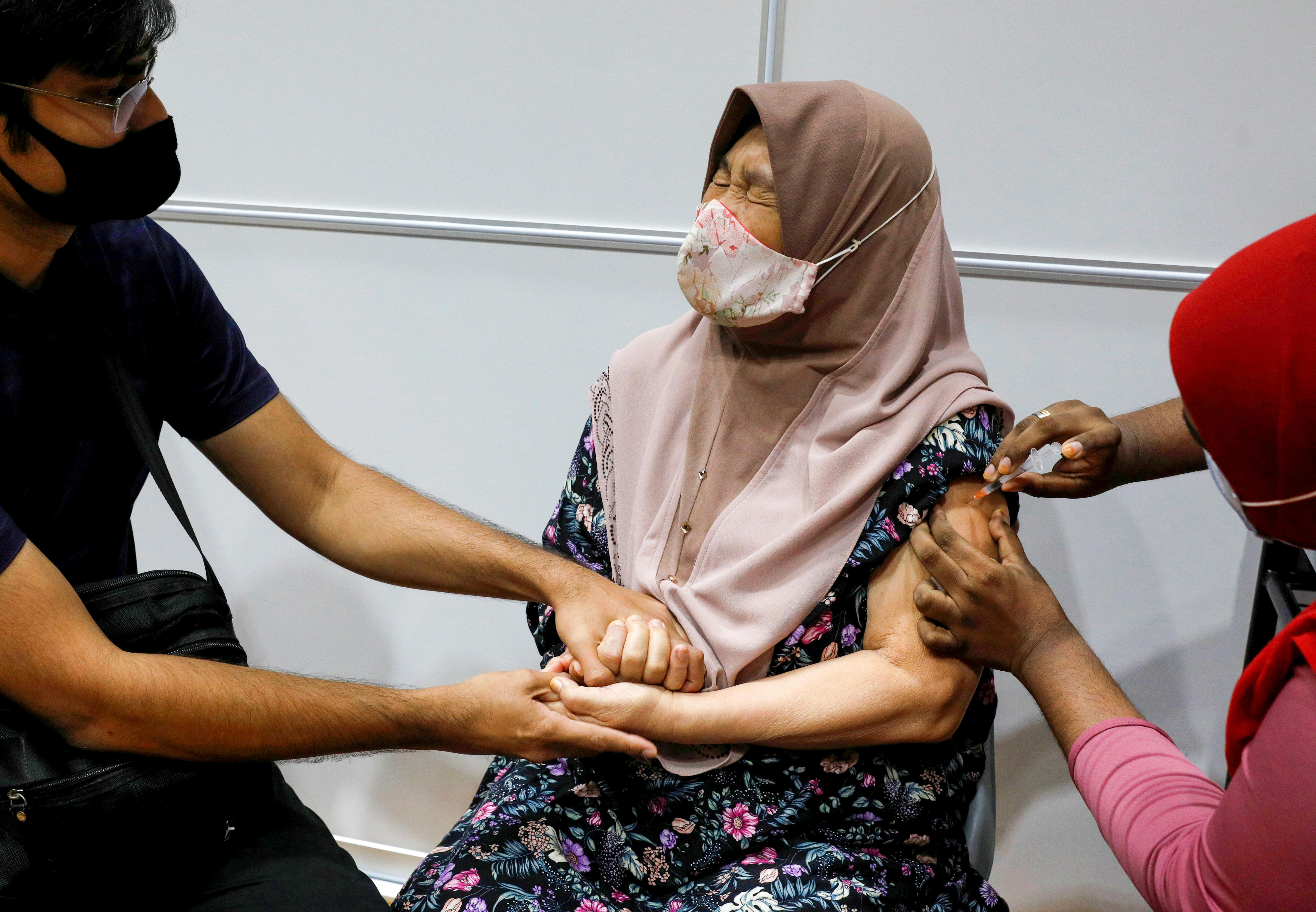 A woman in a mask grimaces and grips a man's hands as a nurse injects her shoulder