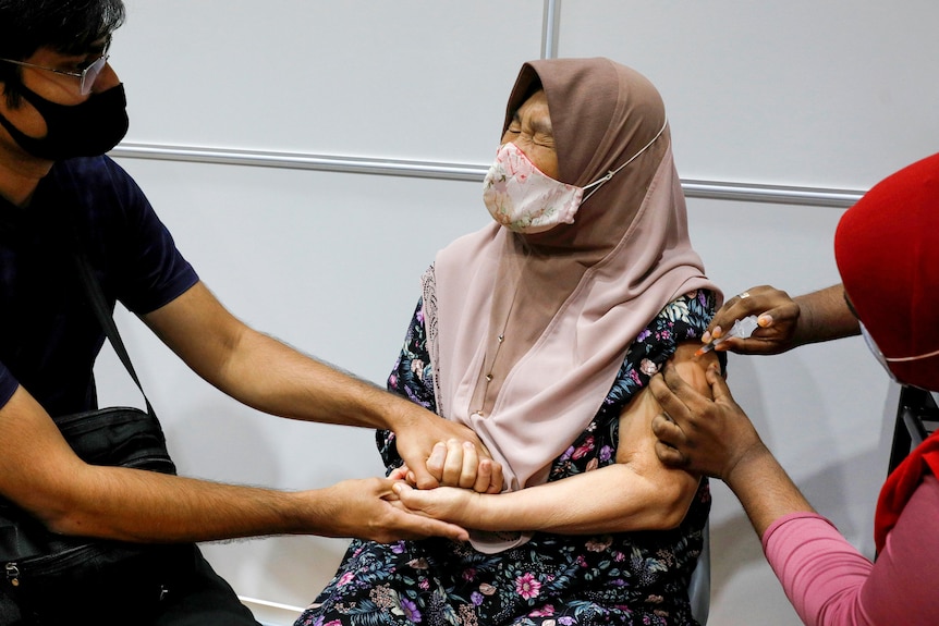 A woman in a mask grimaces and grips a man's hands as a nurse injects her shoulder