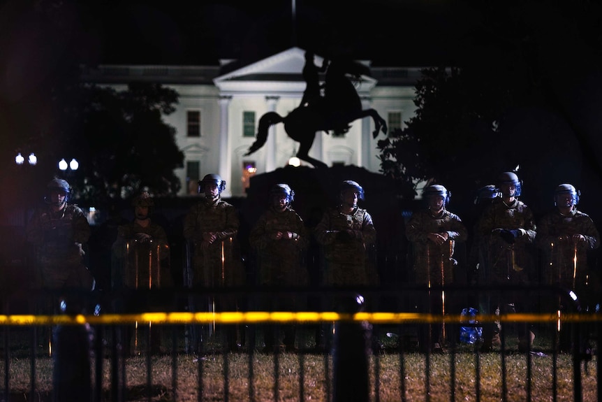 A row of police wearing helmets and stand shoulder to shoulder behind large rectangular shields, with the White House lit up.