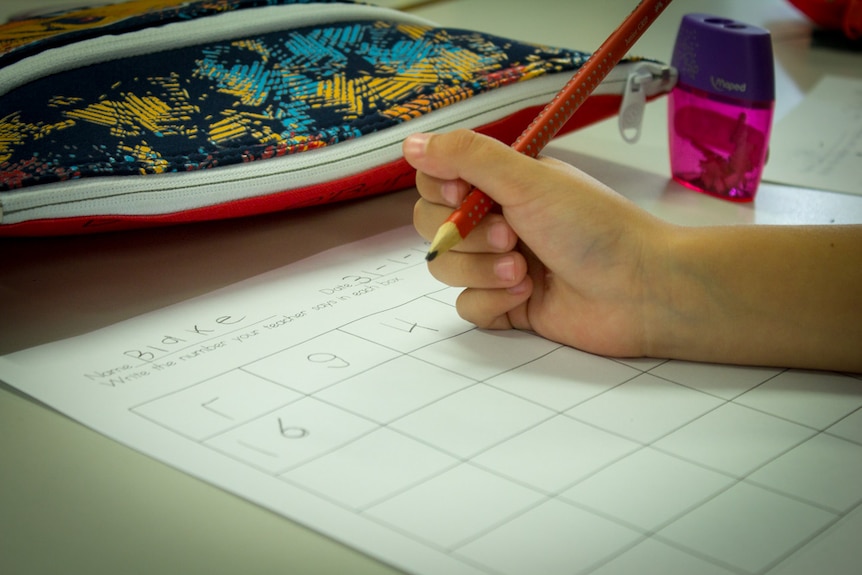 Child holds pencil in hand as he writes letters, pencil case close by.