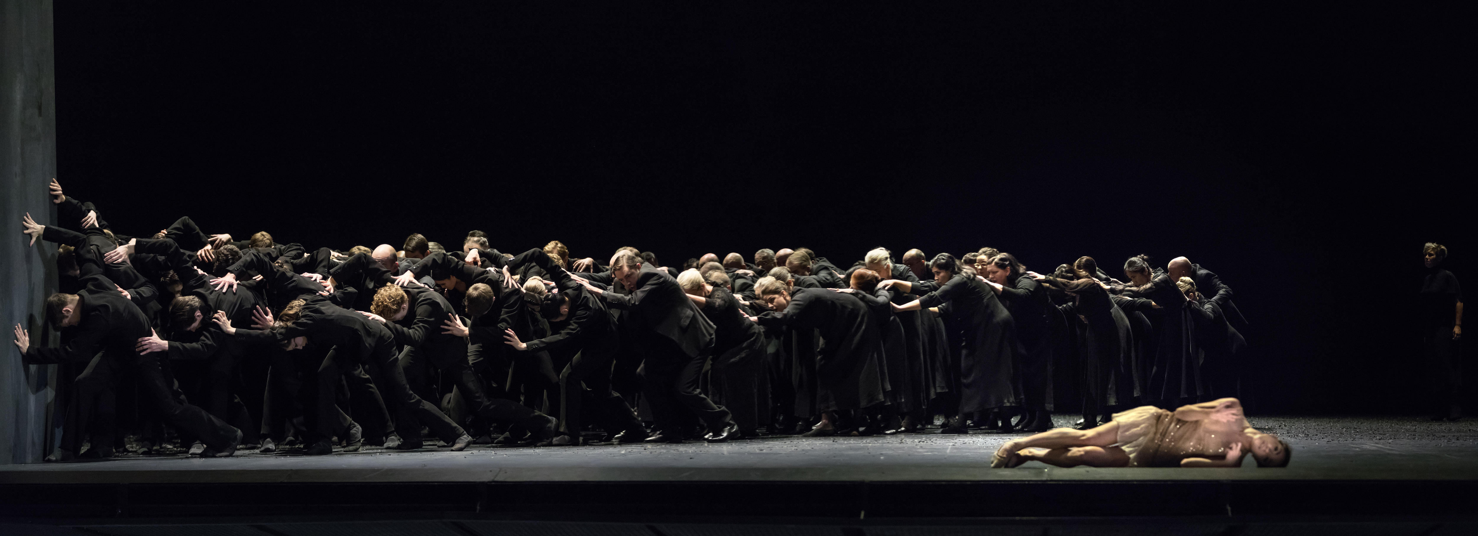 About 50 dancers in black form a chain pushing a wall on a stage with a single dancer in a beige costume lying in front of them
