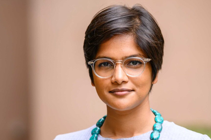 A young Indian woman with short hair, glasses, blue necklace looks seriously, pale background.
