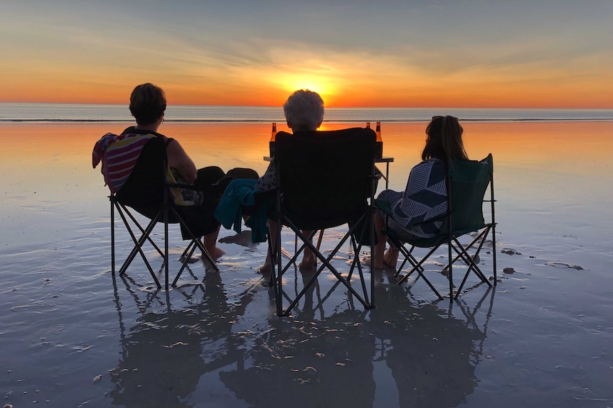 Three people sit in camping chairs on Cable Beach looking out at the ocean and a bright sunset.