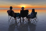 Three people sit in camping chairs on Cable Beach looking out at the ocean and a bright sunset.