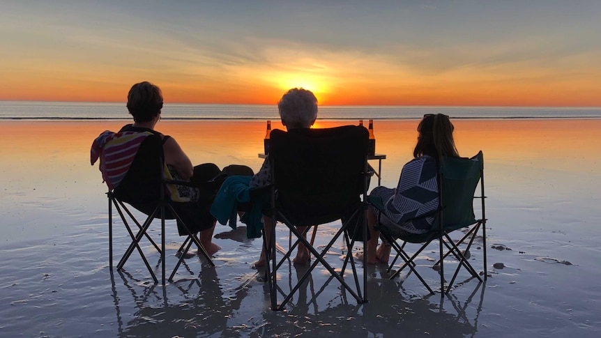 Three people in deckchairs are silhouetted against a setting sun on a deserted beach