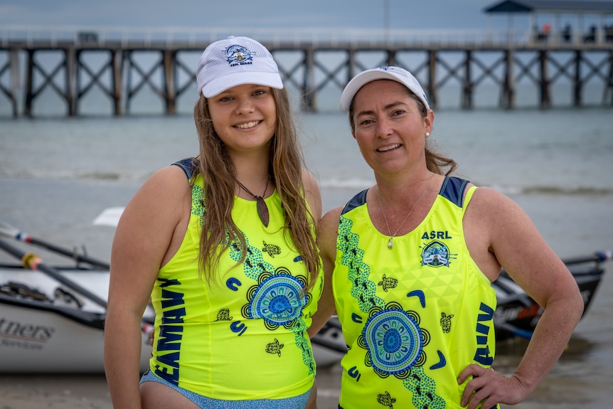 Two women wearing white caps and bright green singlets stand in front of a jetty and ocean