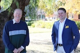 Two men standing standing in front of a tree and a building smiling at the camera.