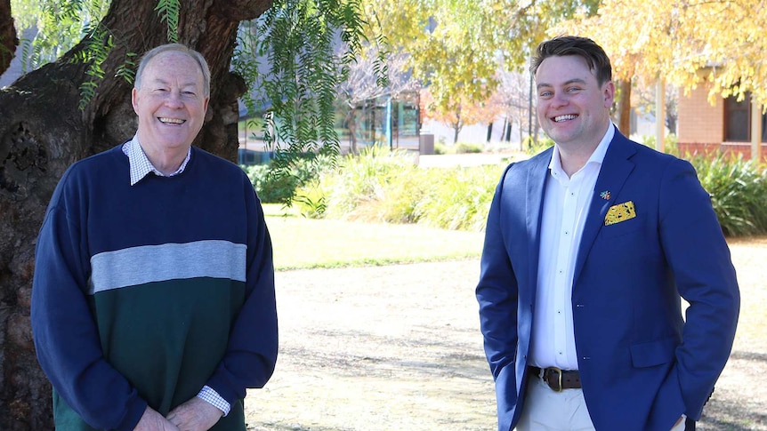 Two men standing standing in front of a tree and a building smiling at the camera.