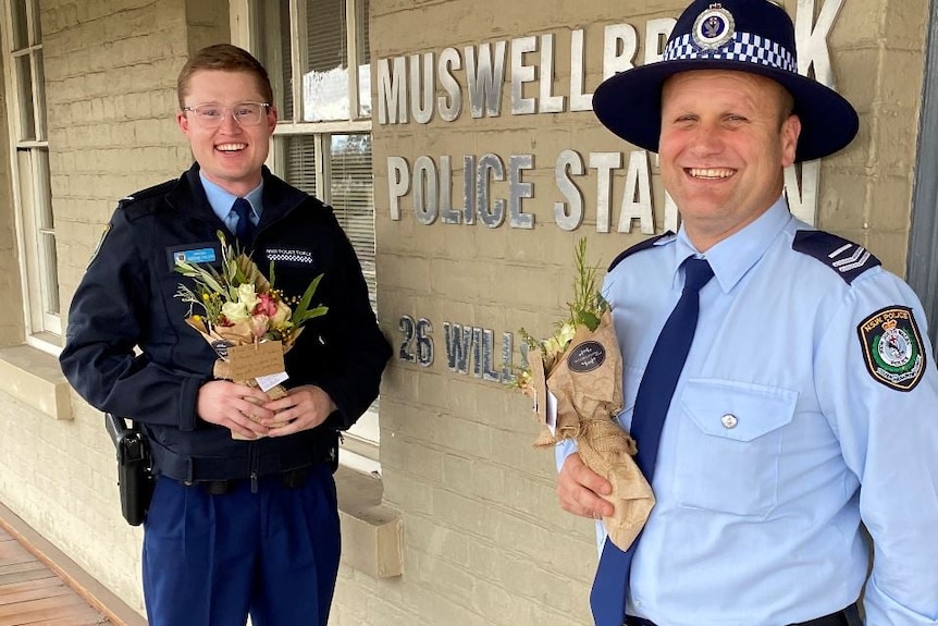 Two male police officers in uniform stand smiling, holding floral posies.