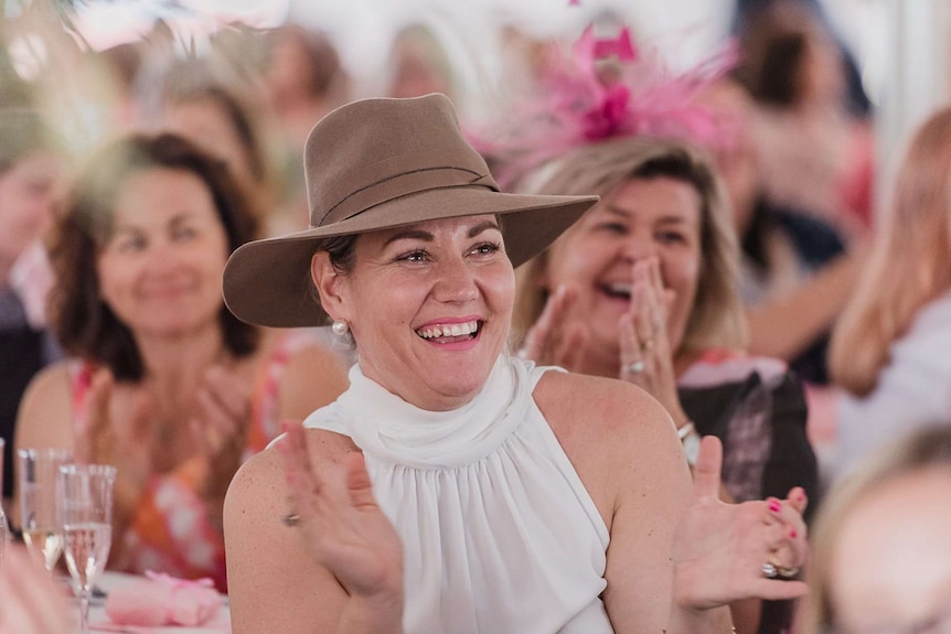 A close up of a woman in a hat applauding, while seated at a table.