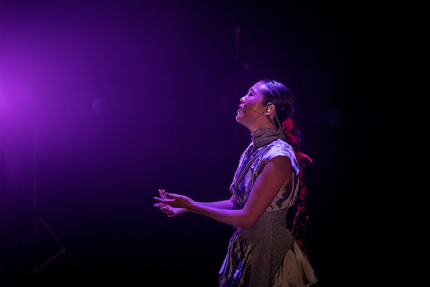 A Chinese Australian in her 30s with long dark hair wears a silver dress and sings under a soft purple light onstage.