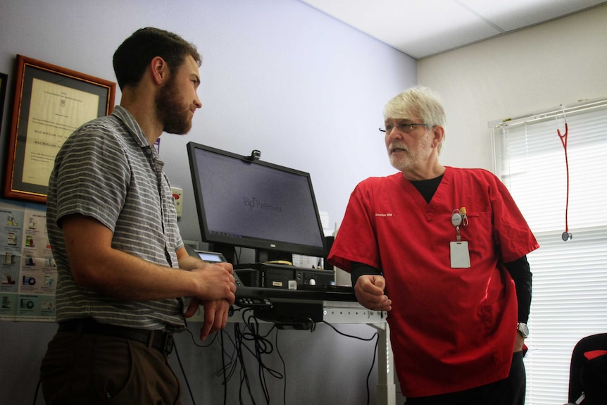 A younger male doctor stands in front of a computer looking at an older male doctor.