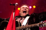 Meat Loaf smiles and holds his arms out wide as he performs with a guitar on stage