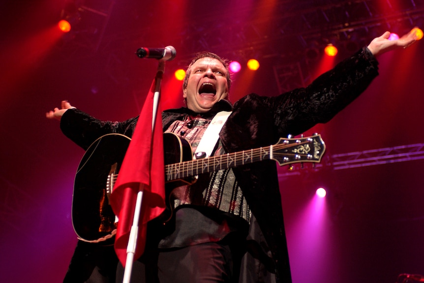Meat Loaf smiles and holds his arms out wide as he performs with a guitar on stage