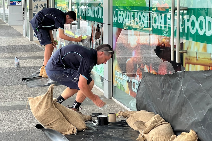 Two men in navy blue workwear taping and sandbagging a shopfront.
