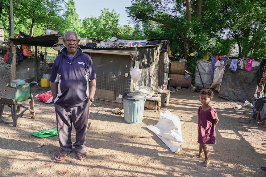 A man and a small child stand in front of a small makeshift hut.