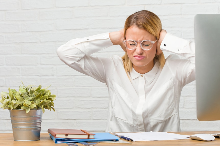 A young woman sits at her desk doing tasks while covering her ears with her hands, angry and tired of distractions.