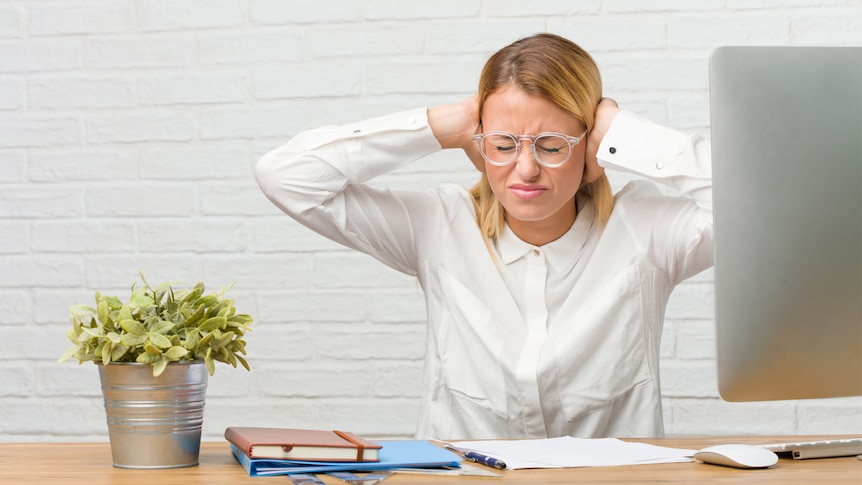 A young woman sits at her desk doing tasks while covering her ears with her hands, angry and tired of distractions.