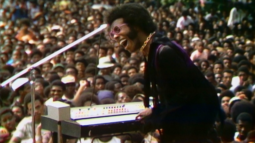 Sly Stone on stage in Summer of Soul wearing a gold chain and purple jacket with the audience in the background.