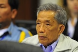 Cambodian man and former Khmer Rouge prison chief Kaing Guek Eav with grey hair sits in a blue shirt and beige jacket.