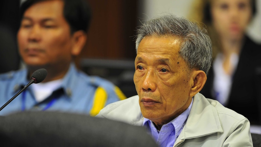 Cambodian man and former Khmer Rouge prison chief Kaing Guek Eav with grey hair sits in a blue shirt and beige jacket.