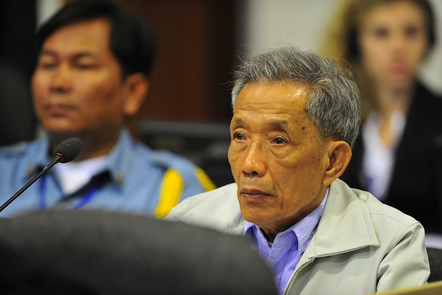 Cambodian man and former Khmer Rouge prison chief Kaing Guek Eav with grey hair sits in a blue shirt and beige jacket.