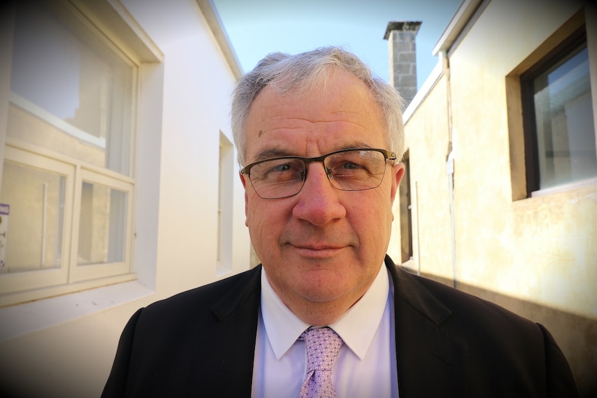 An older, bespectacled man with receding, pale grey hair, formally dressed, looks into the camera lens.