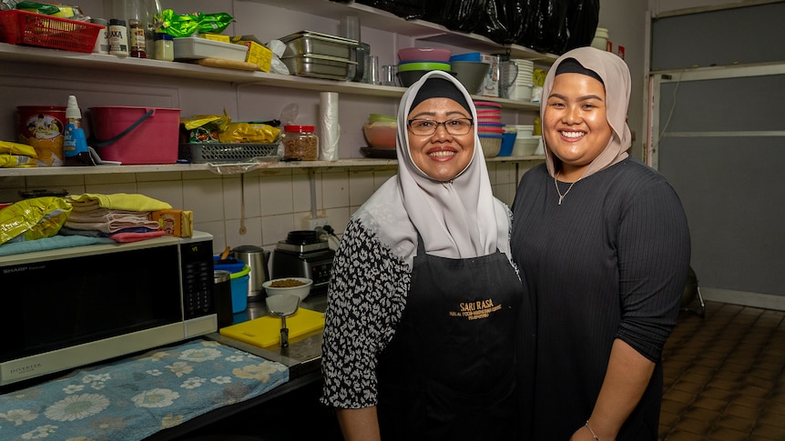 Two women wearing hijabs stand in a restaurant kitchen and are smiling at the camera.
