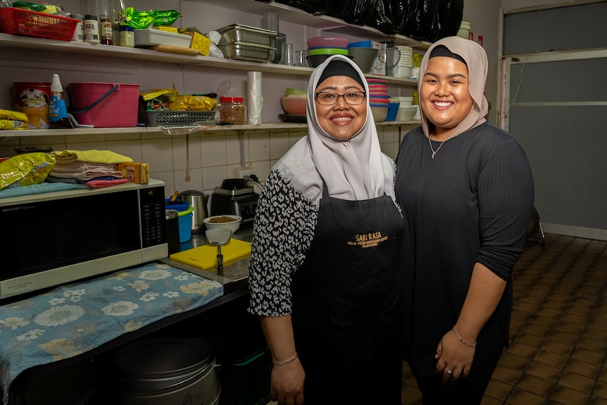 Two women wearing hijabs stand in a restaurant kitchen and are smiling at the camera.