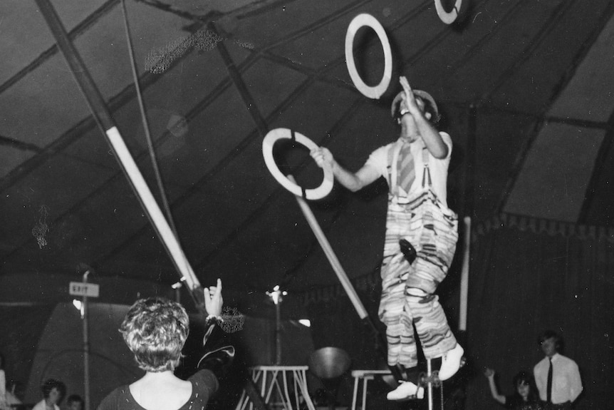 A black and white photo of a unicyclist juggling under a circus big top tent.