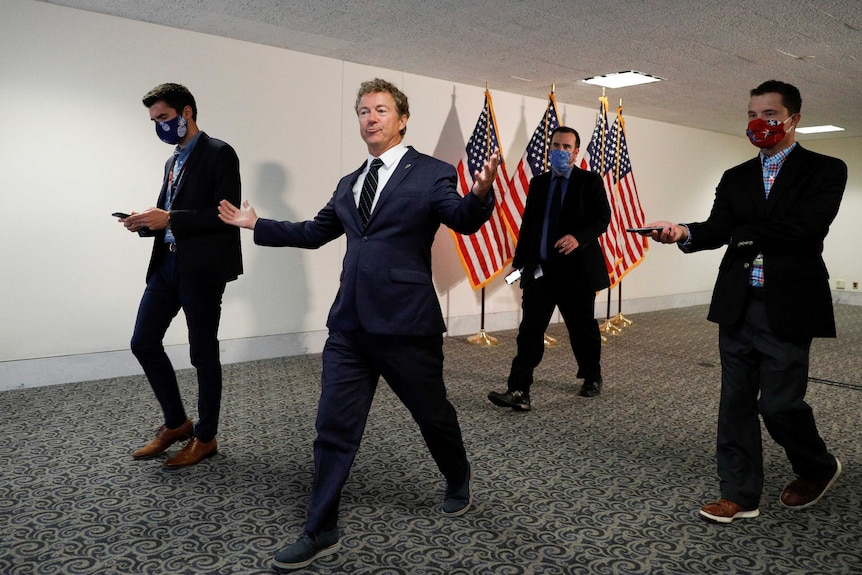 Rand Paul throws his hands up in the air while walking down a hallway surrounded by aides
