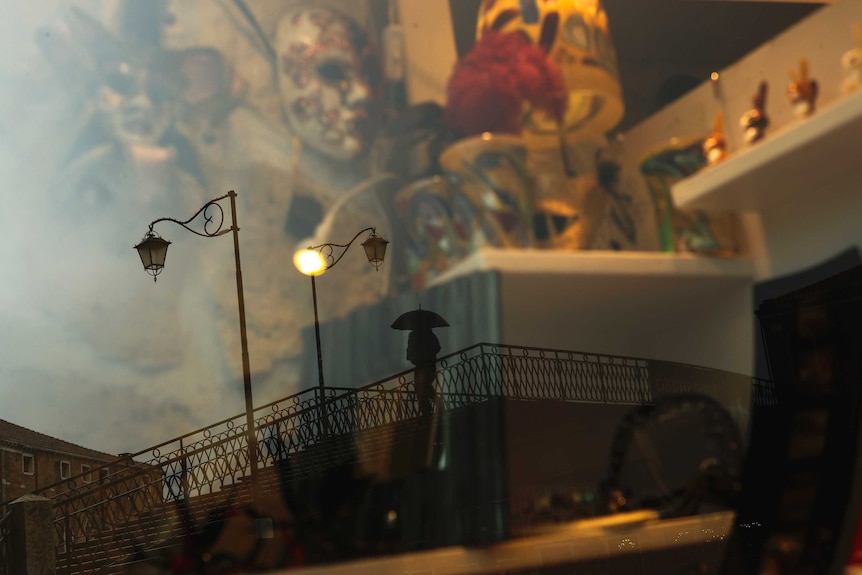 A man, reflected in a carnival masks shop display window, crosses a bridge at the Murano island in Venice.