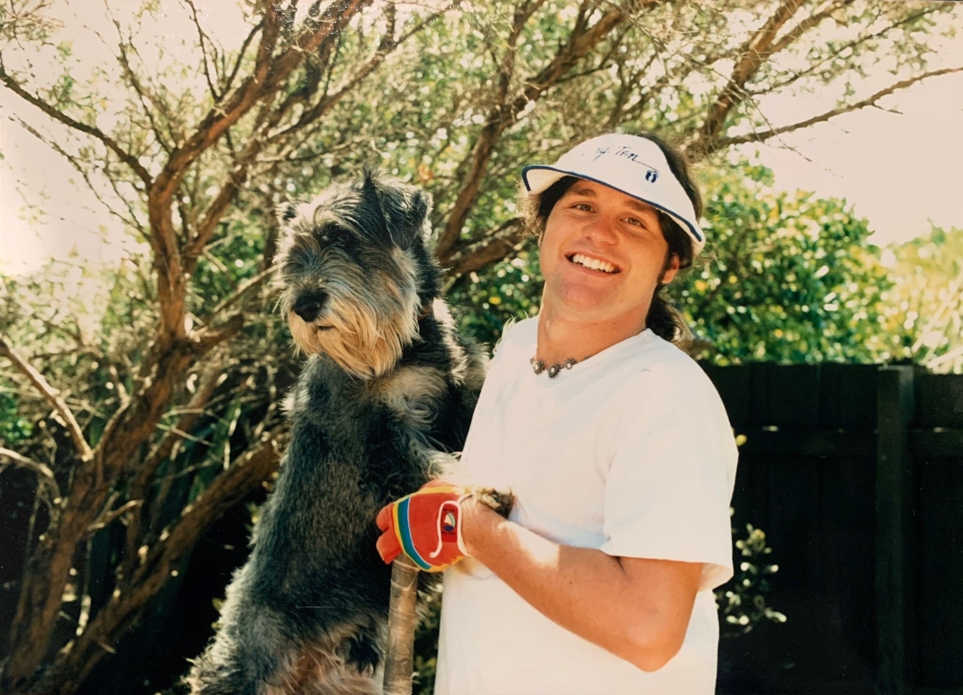 A man in a white shirt and visor smiles and stands next to a grey and white dog.