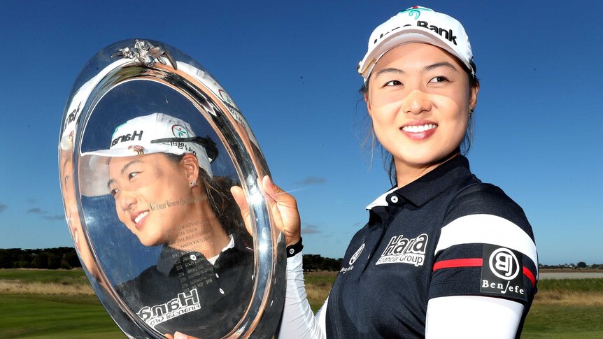 A woman golfer smiles into the camera as her face is reflected in the trophy she holds.