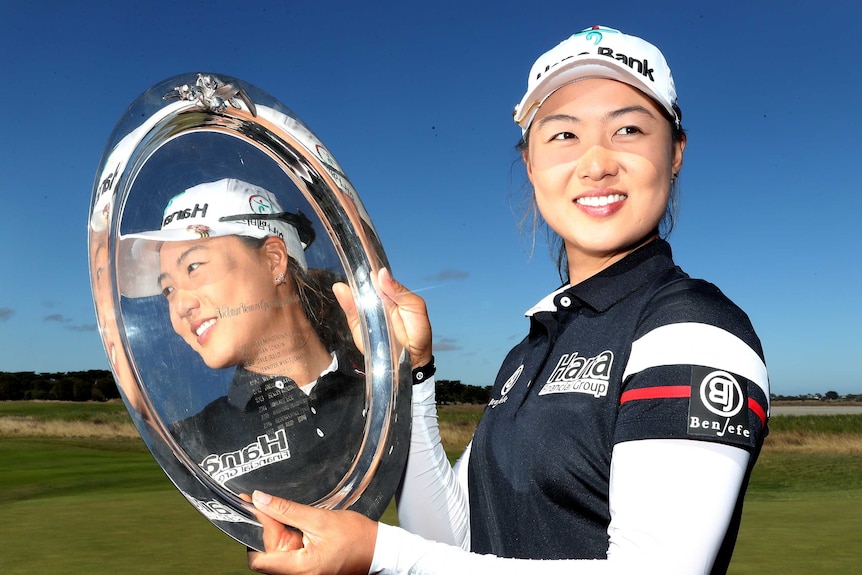 A woman golfer smiles into the camera as her face is reflected in the trophy she holds.