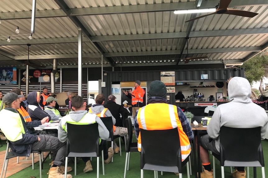 A group of men wearing hi vis clothes listening to a talk presentation.