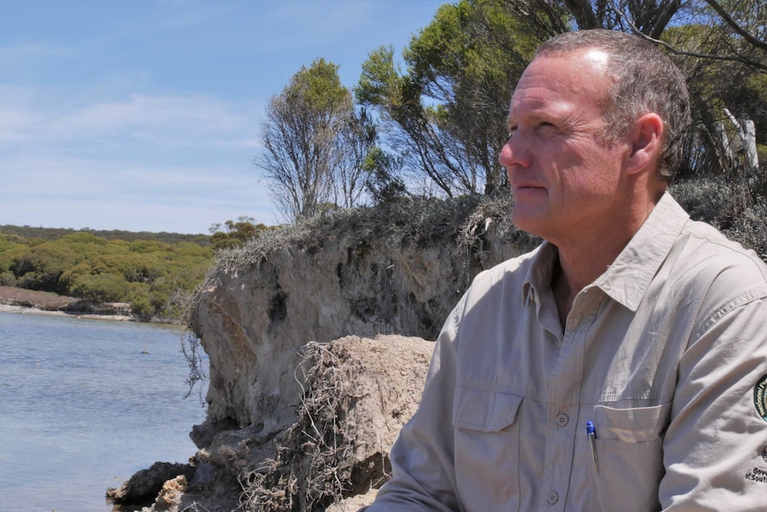 A man with short hair sits on the banks for a small lake.