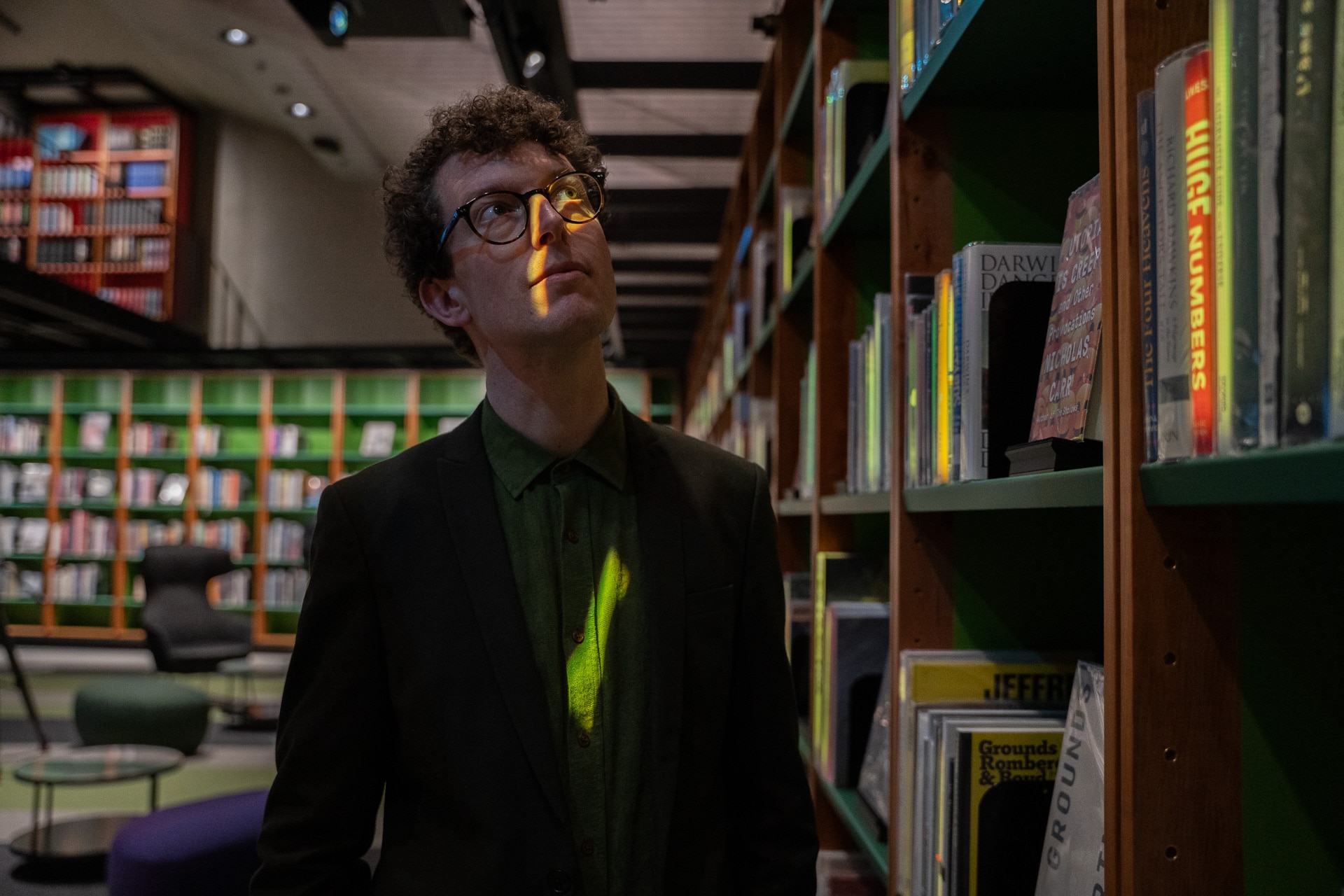 Nic Whyte gazes up at the bookshelves in a library. Colours light up the books and hit face.