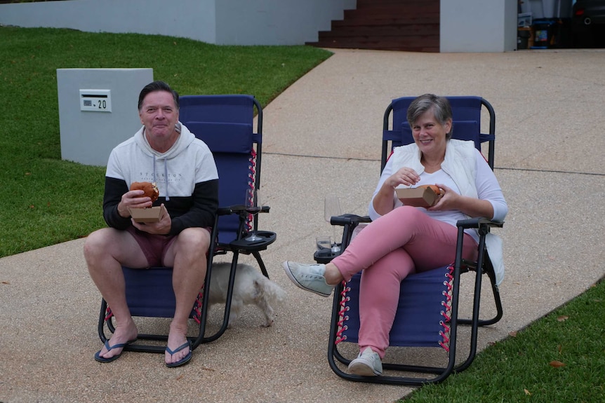 A man and woman sit on chairs on their driveway eating burgers.