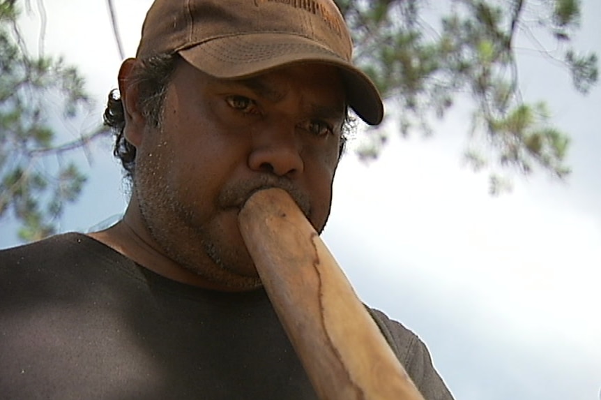 A man wearing a brown cap and shirt plays the didgeridoo.